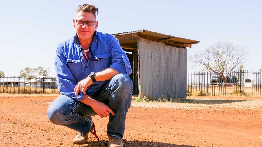 A middle aged man squats on red dirt in a rural area.