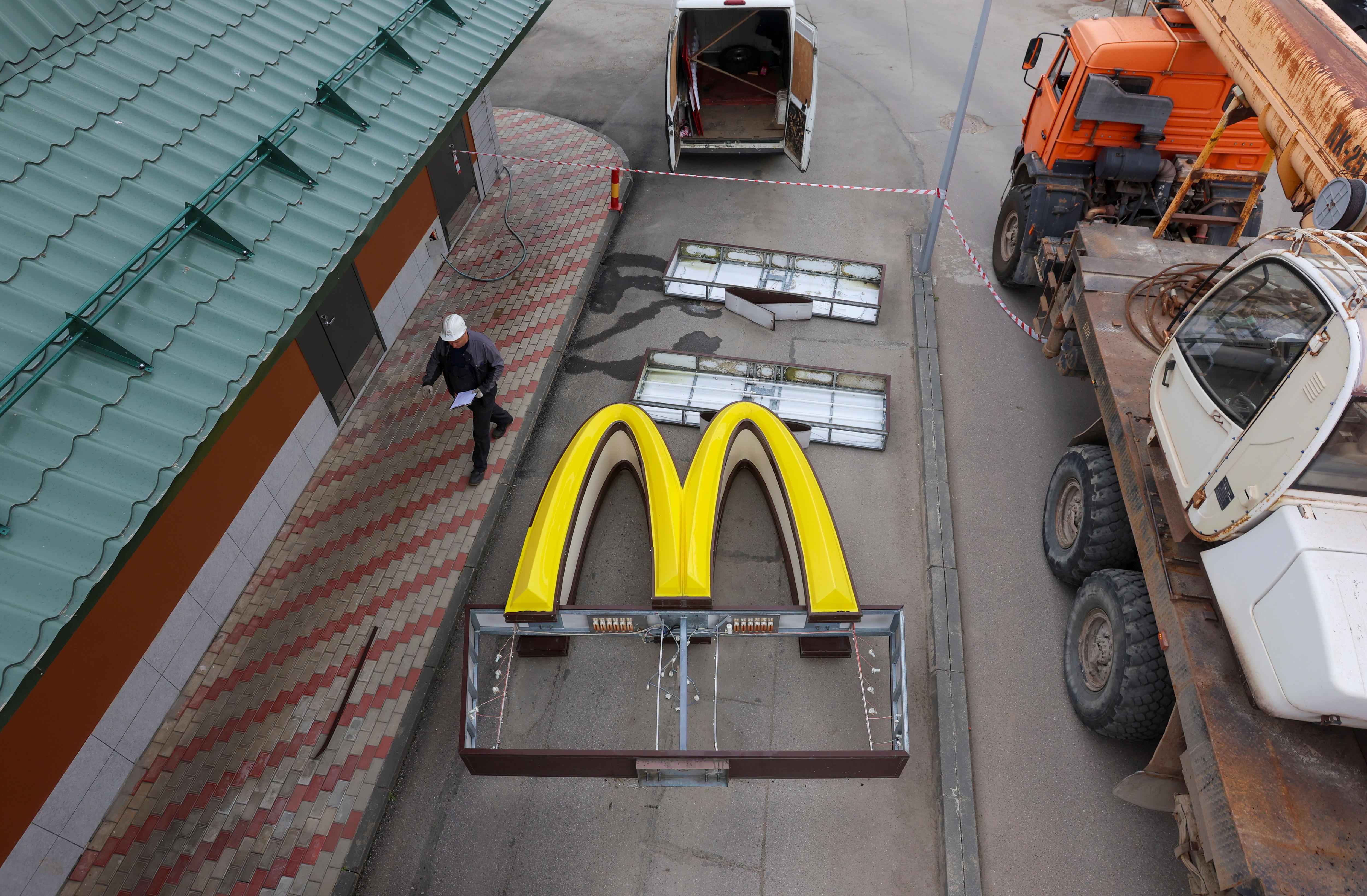 An aerial shot shows a construction worker wearing a hard hat walking past a giant golden M on the ground