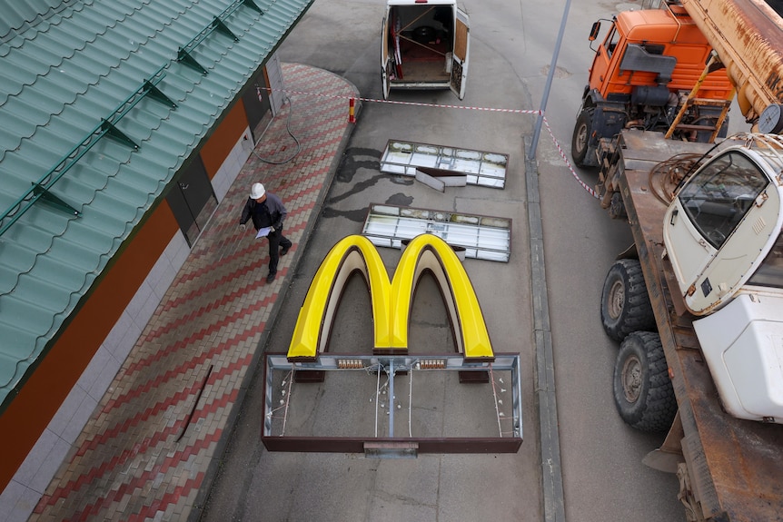 An aerial shot shows a construction worker wearing a hard hat walking past a giant golden M on the ground