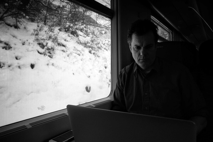 Black and white picture of Cannane sitting by train window with snow outside looking at laptop.