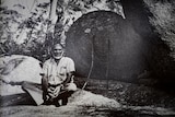 Black and white photo of an Aboriginal elder sitting cross-legged on a large rock in the forest