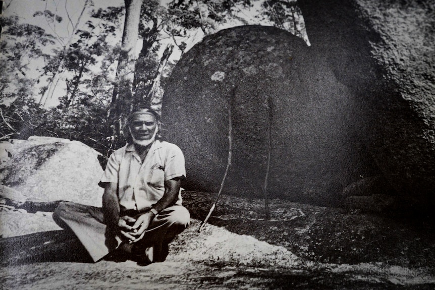 Black and white photo of an Aboriginal elder sitting cross-legged on a large rock in the forest