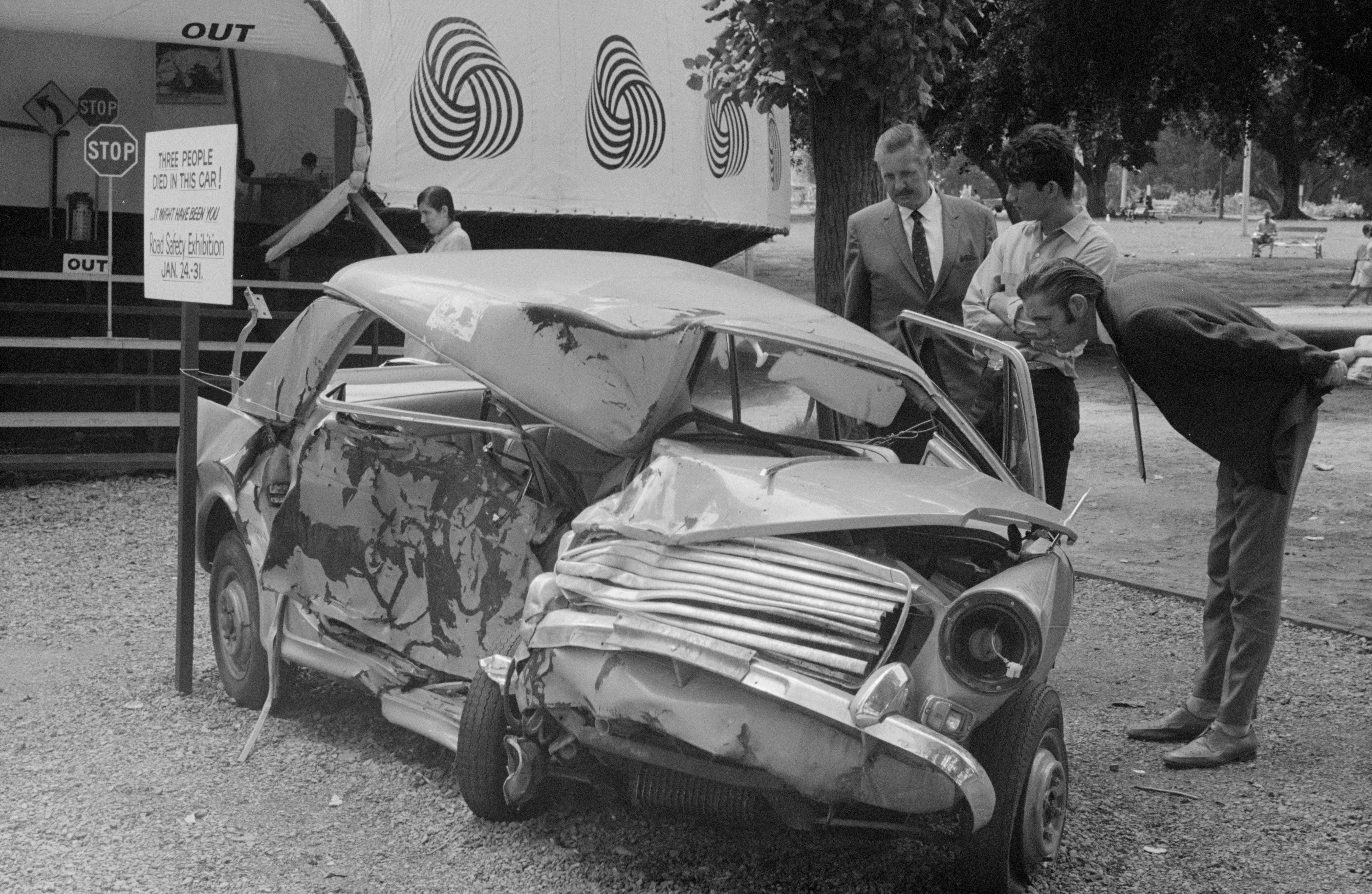 A 1960s black and white photo of a severely smashed car with three men looking inside