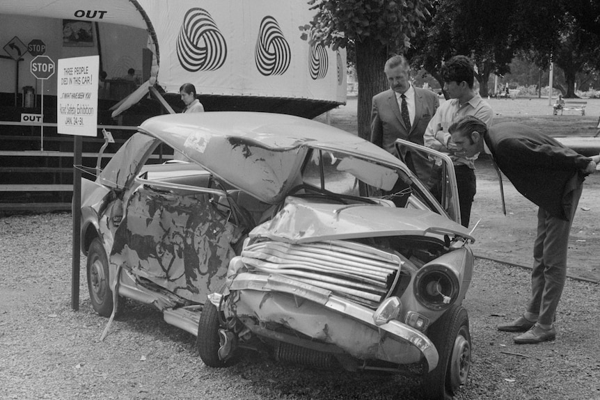 A 1960s black and white photo of a severely smashed car with three men looking inside