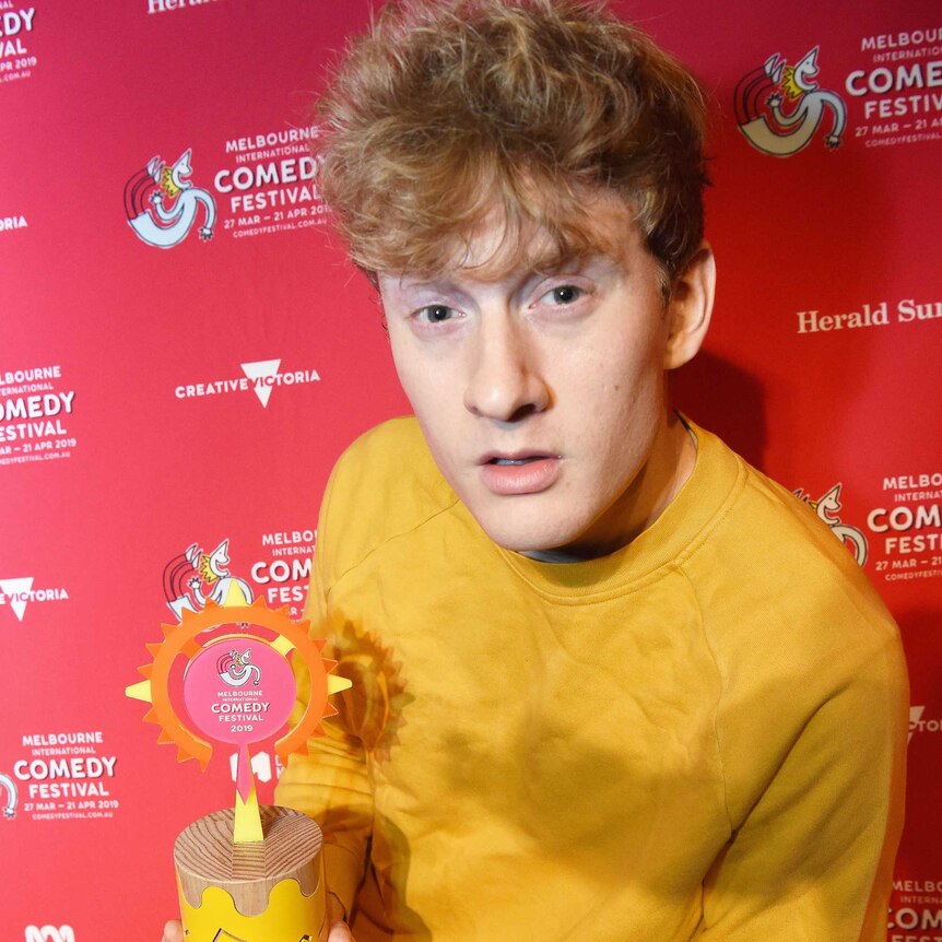 Comedian James Acaster with a deadpan expression holding the MICF Award trophy.