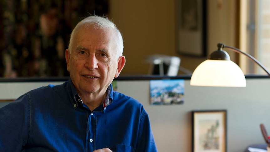 Off centre portrait of older Caucasian man with bright blue collared shirt smiling, in his study.