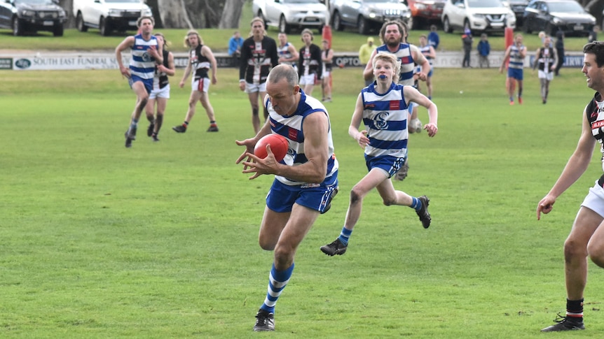 A man catches a football on the run, players behind him on a green oval