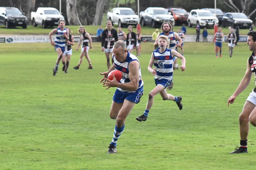 A man catches a football on the run, players behind him on a green oval