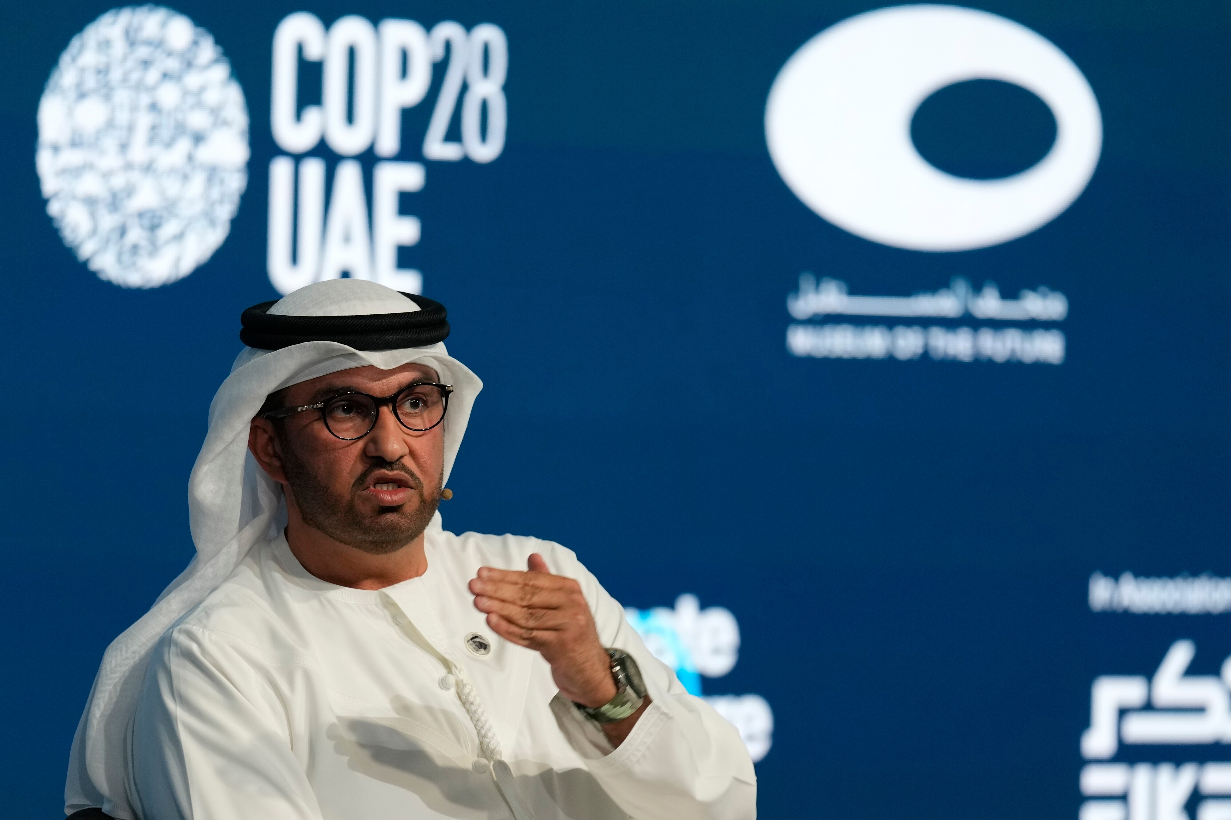 A man wearing a Ghutra sits and speaks in front of a backdrop with the 'COP28UAE' logo