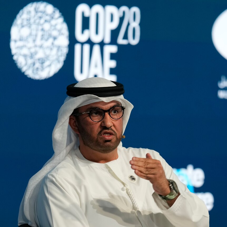 A man wearing a Ghutra sits and speaks in front of a backdrop with the 'COP28UAE' logo