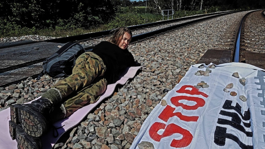 A woman lays on a a train train with a sign that reds ' stop the machine'.