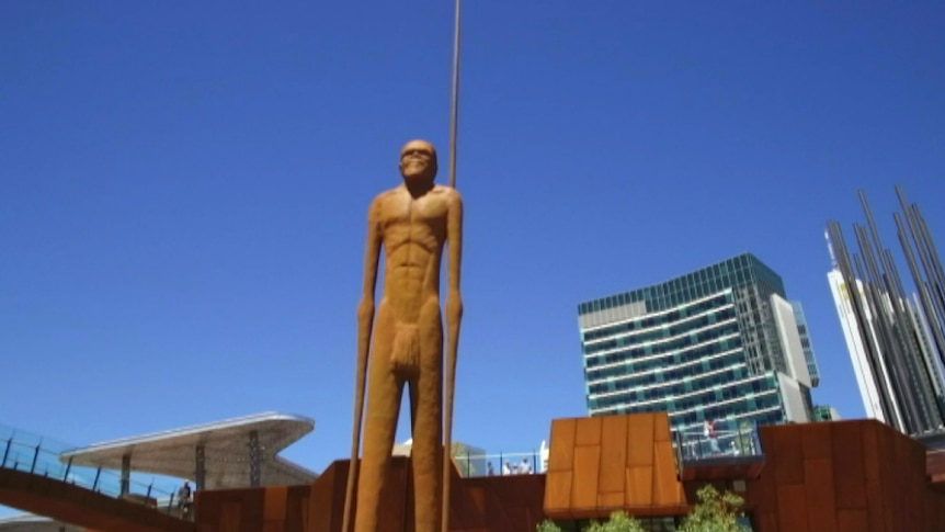 A tall statue of Yagan stands in front of a blue sky and the Perth city skyline.