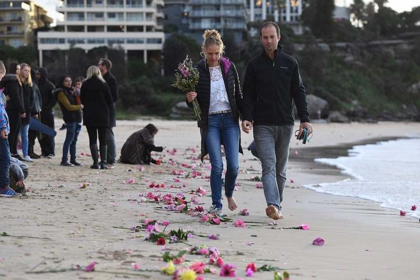 A couple walk down the beach holding hands while the woman holds a bunch of flowers.