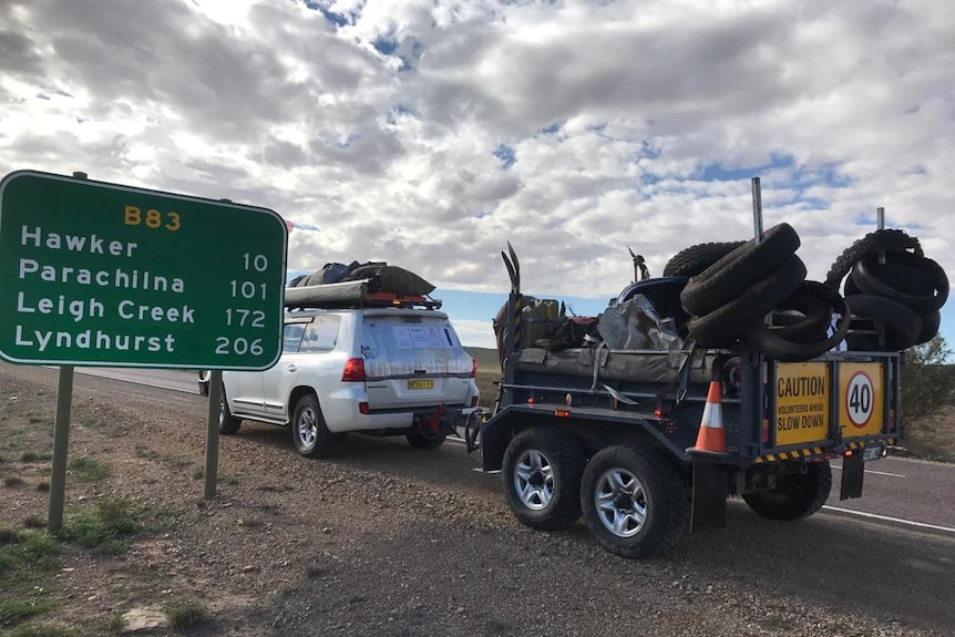 A white four-wheel-drive pulling a trailer load of tyres passes a sign that lists towns between Quorn and Lyndhurst.