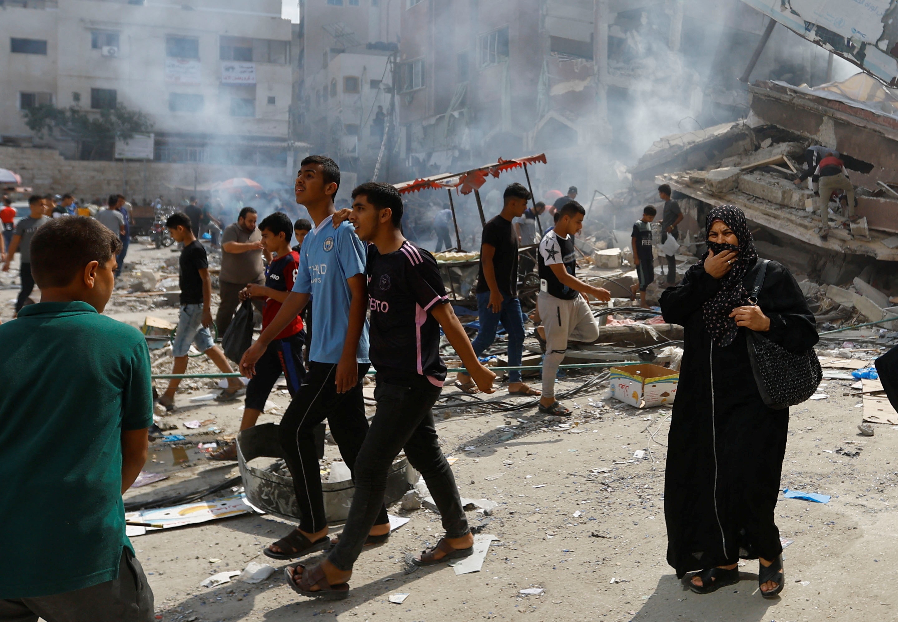 Young Palestinian men and a middle-aged woman walk past smoking concrete wreckage on a city street.