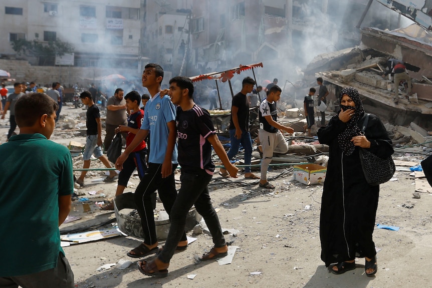 Young Palestinian men and a middle-aged woman walk past smoking concrete wreckage on a city street.