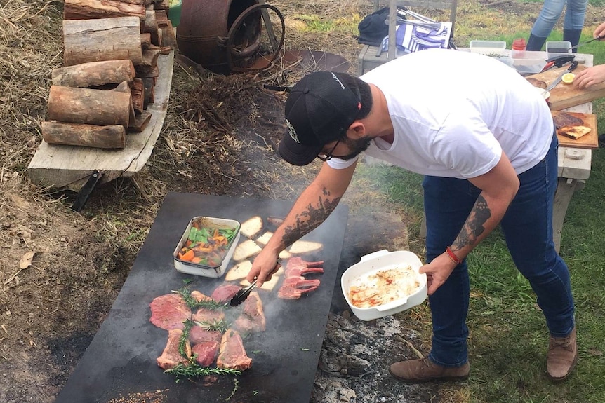 A man in jeans, a white t-shirt and black cap leans over a cooking plate over a fire.