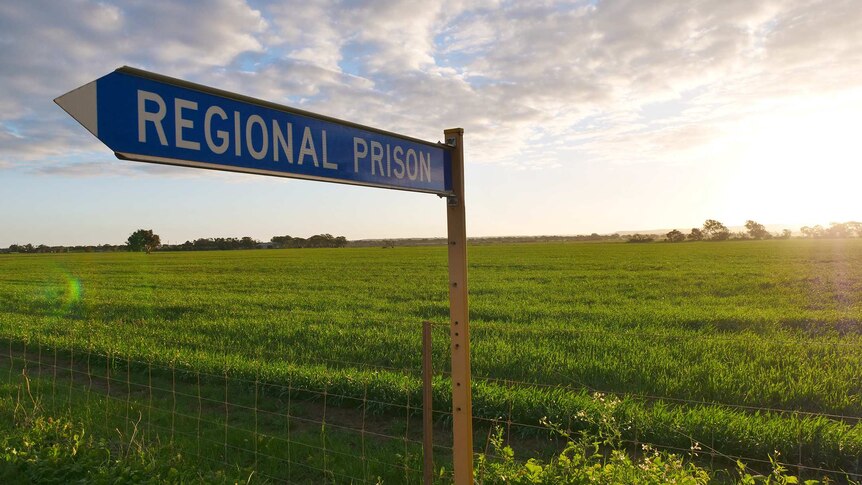 A blue road sign points the way to Greenough Regional Prison in front of a lush green field.