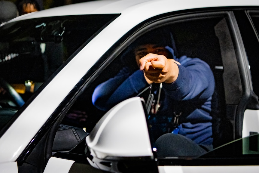 A man sitting in the driver's seat of a car at night holds up his arms in a gesture towards the camera.