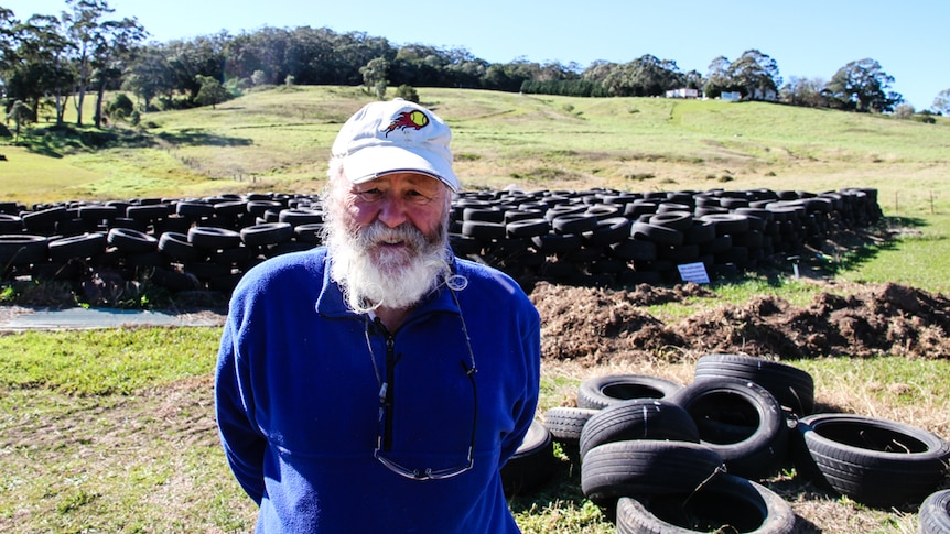 A beared man looks at the camera. Behind him is a maze made of car tyres.