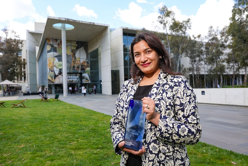 A woman smiles holding a blue rectangular trophy in front of a modern building.