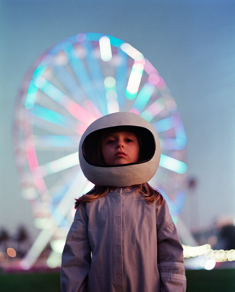 A young girl wearing a spacesuit costume and helmet standing in front of a pink and blue Ferris wheel