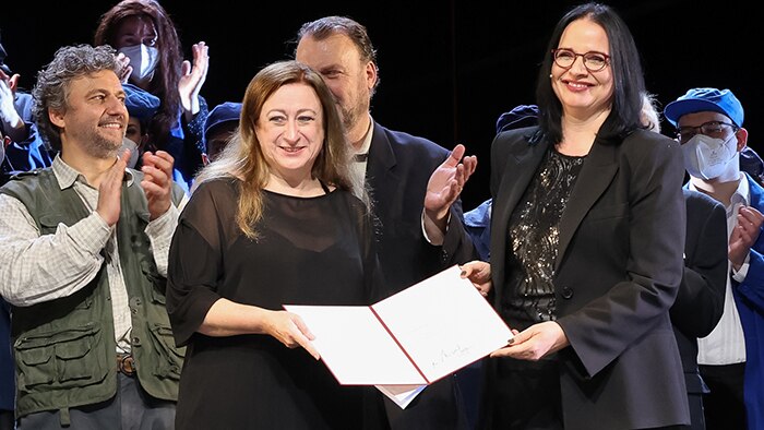 Simone Young holds an award with Andrea Mayer in front of a clapping opera chorus.