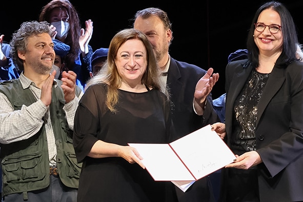 Simone Young holds an award with Andrea Mayer in front of a clapping opera chorus.
