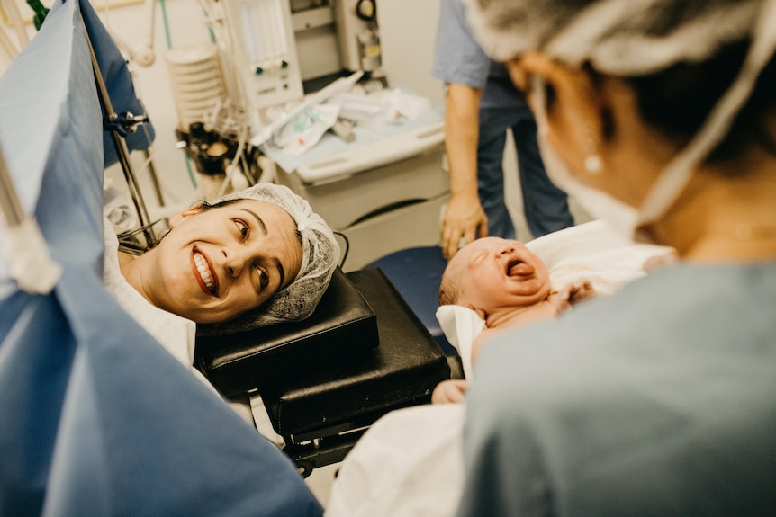 a woman on an operating table smiles at a newborn baby held by a nurse.