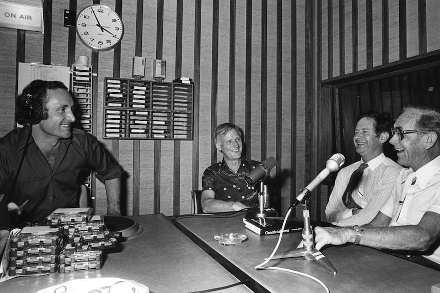 Black and white photo of Williams in radio studio behind the mic talking to three male guests.