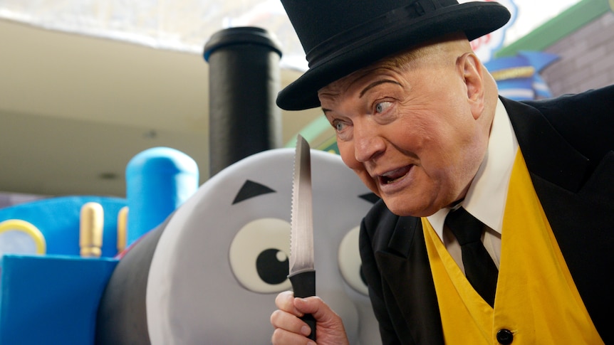 Bert Newton with a yellow vest and black hat looks curiously next to Thomas the Tank Engine