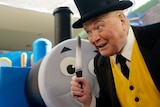Bert Newton with a yellow vest and black hat looks curiously next to Thomas the Tank Engine