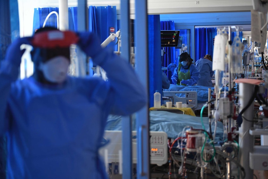 A healthcare worker wearing full PPE takes off a face shield while in the background other workers tend to a patient