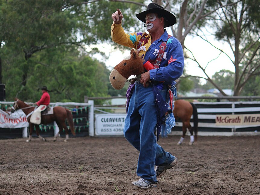 A rodeo clown with colourful clothes and facepaint rides a toy horse across rodeo ground.