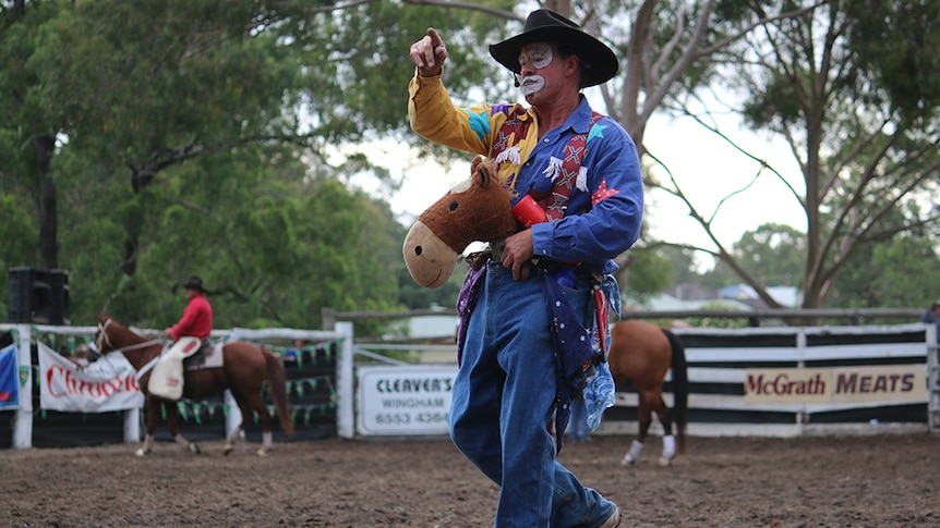 A rodeo clown with colourful clothes and facepaint rides a toy horse across rodeo ground.