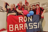 A group of people hold up a sign saying 'Ron Barassi' wearing Demons colours of red and blue.