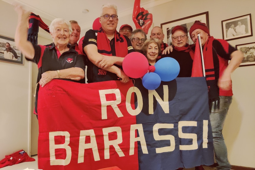 A group of people hold up a sign saying 'Ron Barassi' wearing Demons colours of red and blue.