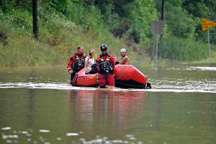 Dos personas se sientan dentro de un bote inflable rojo mientras dos rescatistas caminan por las aguas de la inundación.