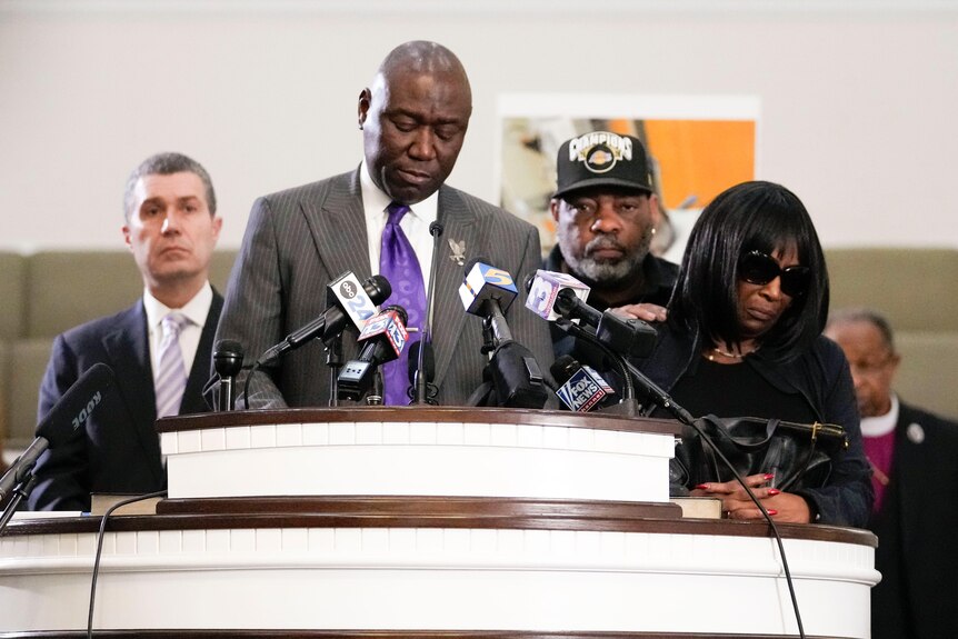 A man stands in front of microphones at a press conference, a sad couple stands next to him. 