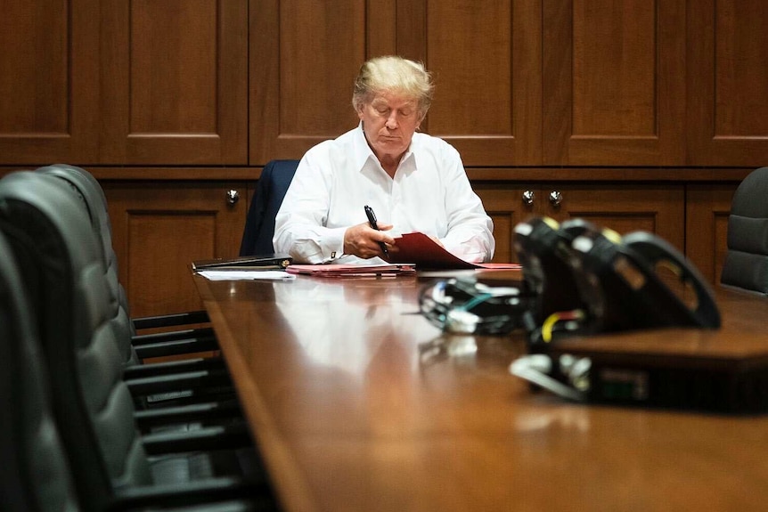 In a white business shirt, Donald Trump looks at some files while sitting at an otherwise empty boardroom desk