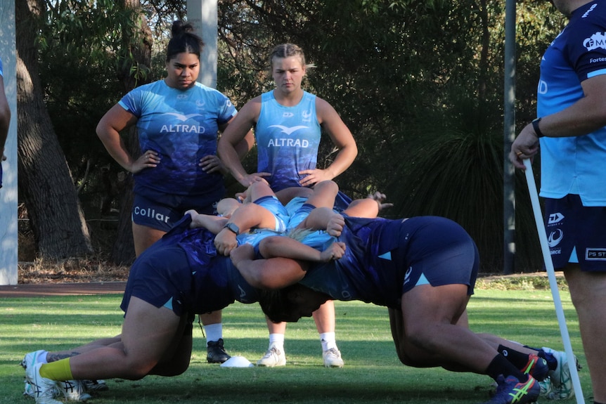 Female rugby players simulate a small scrum as two other women watch on.