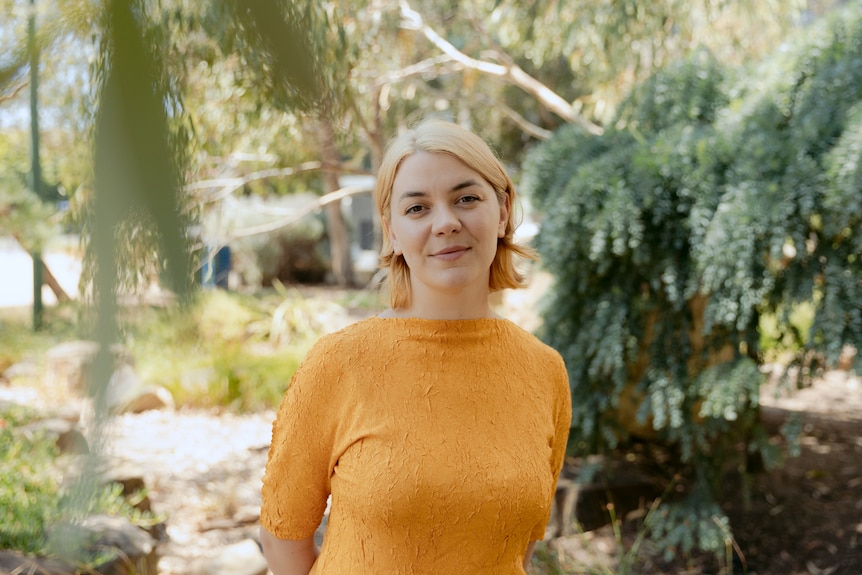 A Wiradjuri woman with light blonde hair and brown eyes wears a yellow crepe dress and stands among greenery in a park.