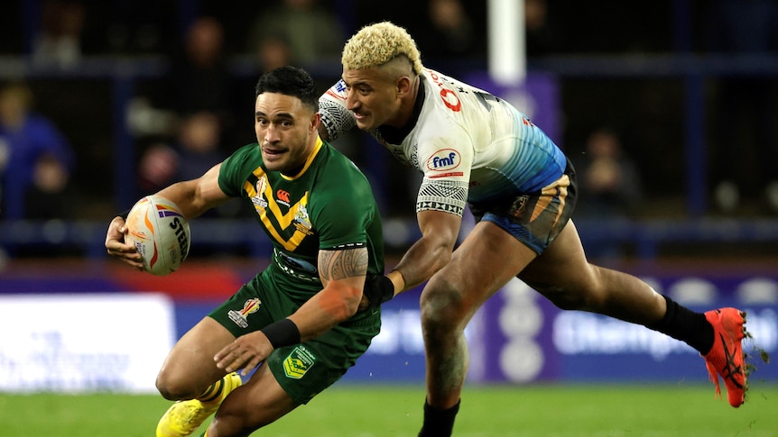 Australia's Valentine Holmes holds a football and runs as a Fijian player leans to tackle him from behind. 