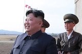 Kim Jong-un looks at what appears to be an airfield, as two military officials stand behind him. He is smiling.