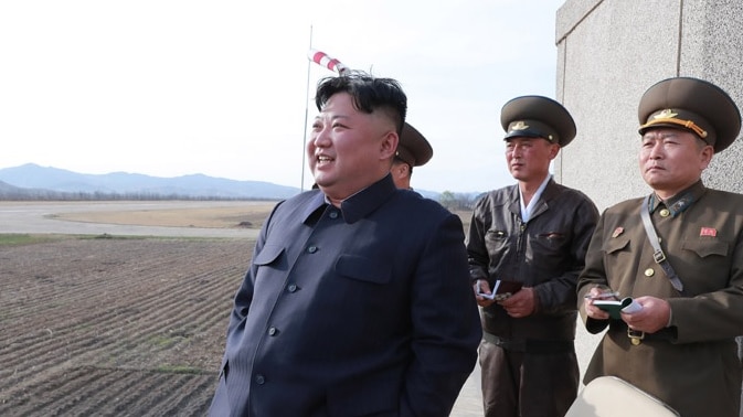 Kim Jong-un looks at what appears to be an airfield, as two military officials stand behind him. He is smiling.