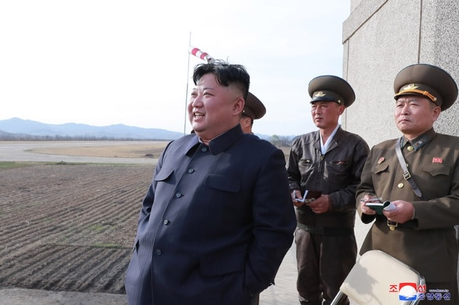 Kim Jong-un looks at what appears to be an airfield, as two military officials stand behind him. He is smiling.