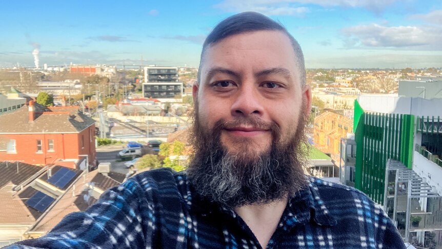 A middle-aged man with a beard takes a selfie on the balcony of his new apartment, with the city in the background.