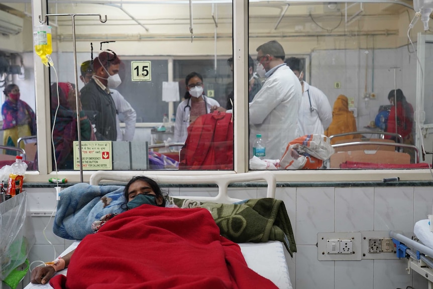 A young Indian woman in a face mask covered in a red blanket lies in a hospital bed