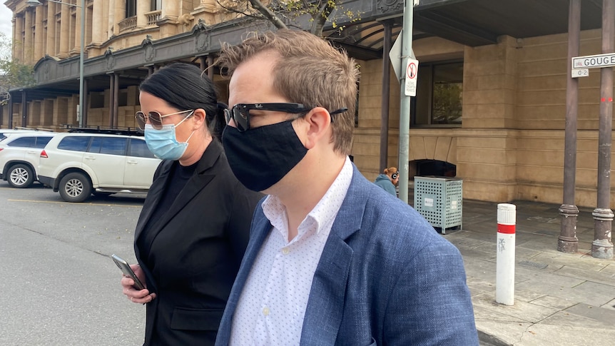 Side profile of a woman and man, both wearing sunglasses and face masks, walking. Behind them is the court building