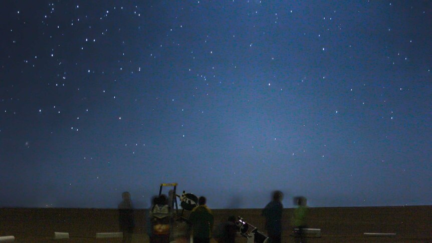 A blue starry sky, with some blurred people looking into large, two metre telescopes.